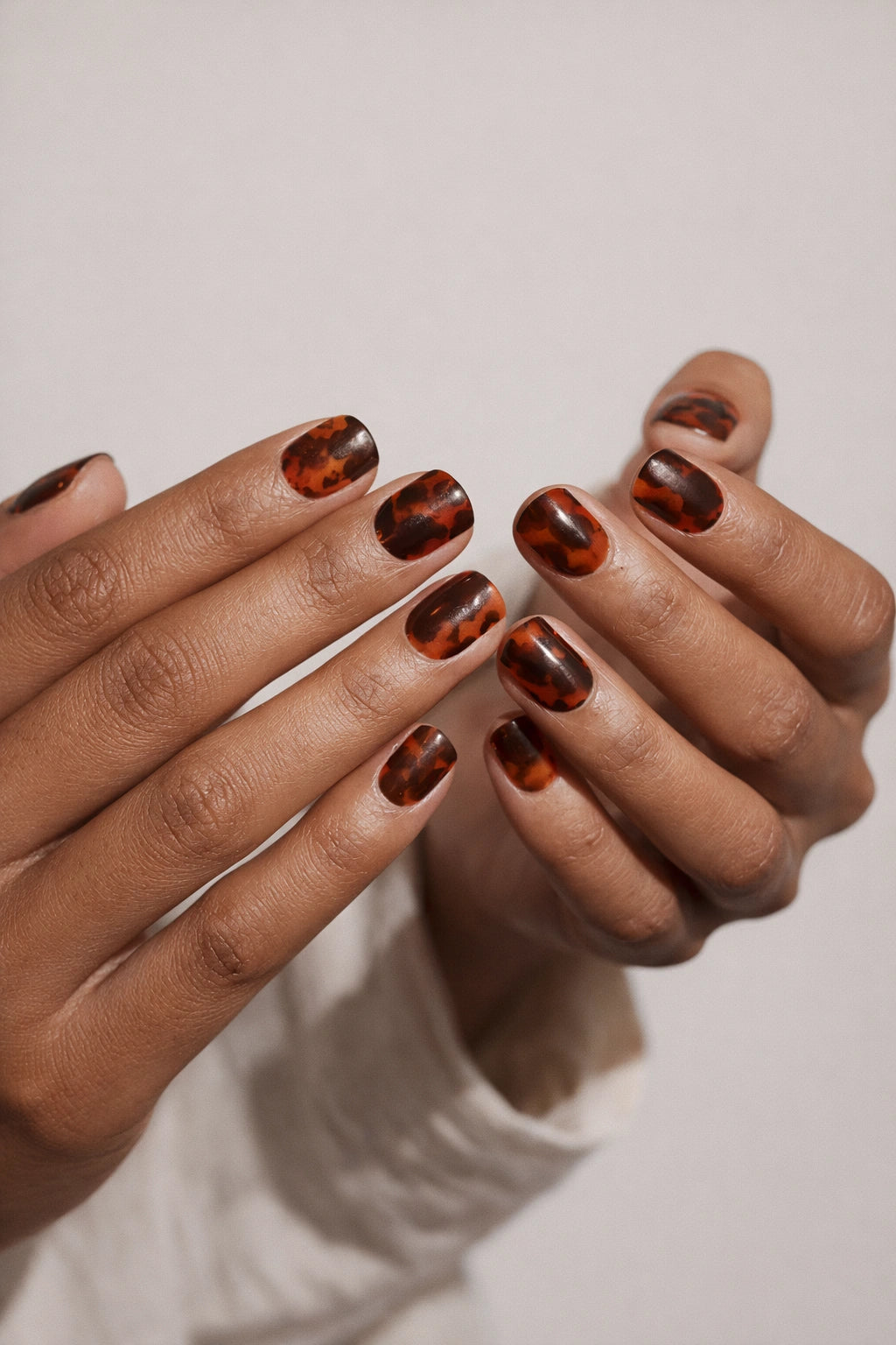 Tortoiseshell semi-cured gel nail stickers, featuring a warm amber and deep brown semi-transparent glossy finish on medium-deep brown skin, photographed under soft studio lighting with a white textured long-sleeve outfit.