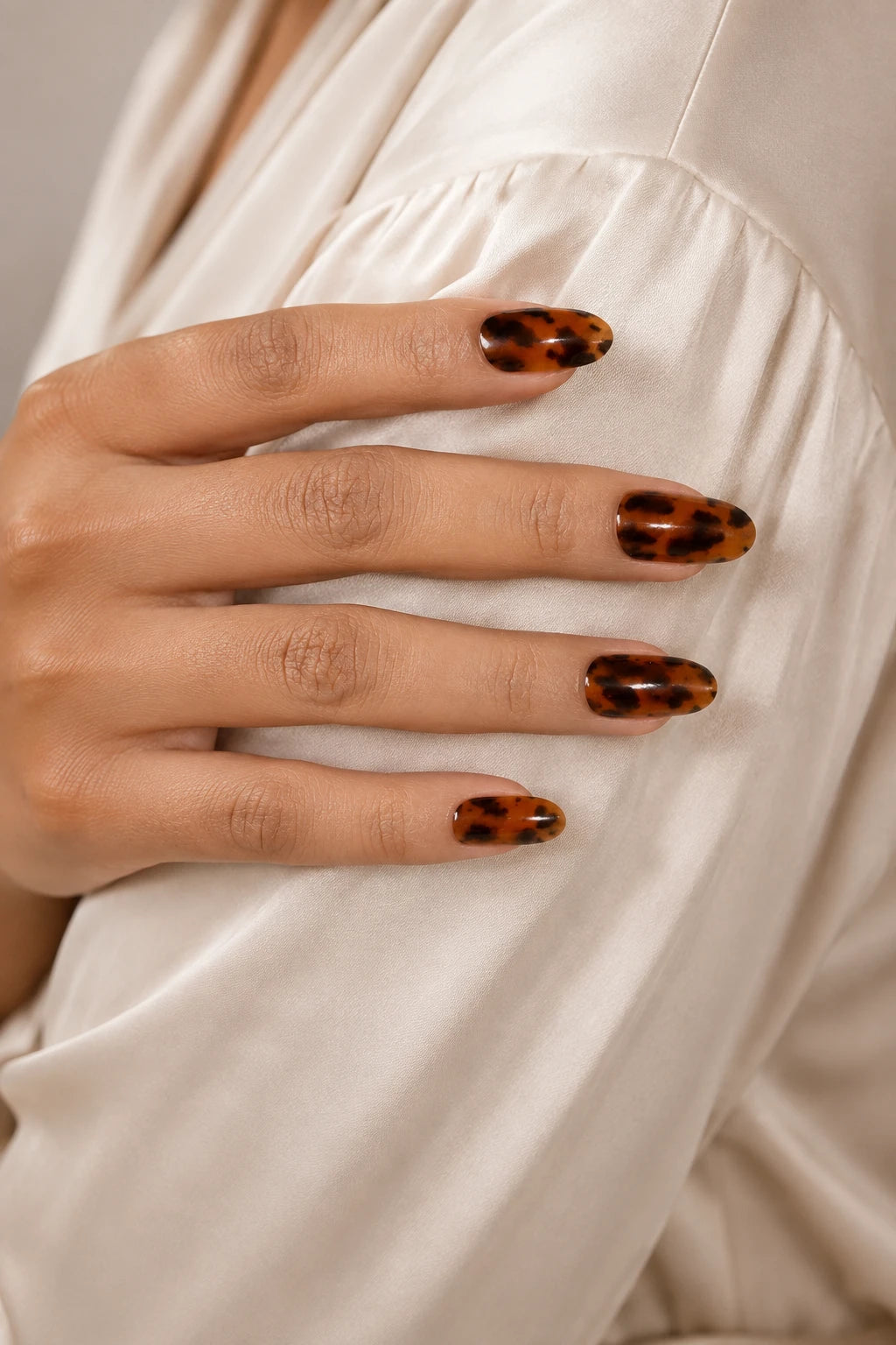 UV-free gel nail wraps featuring a glossy tortoiseshell pattern in warm amber and black, shown on medium-brown skin against soft cream satin.