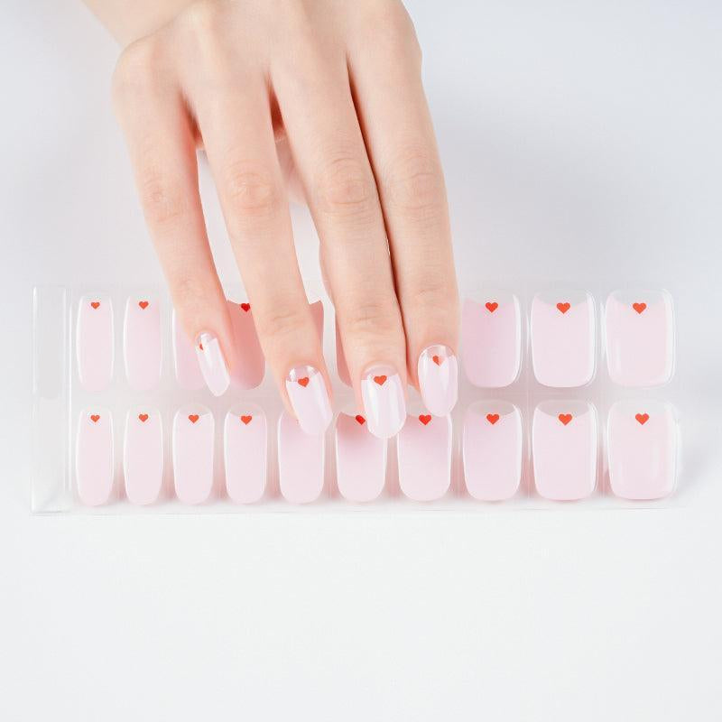 Hand holding sheet of pastel pink gel nail stickers with small red heart designs.