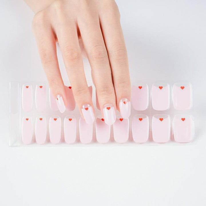 Hand holding sheet of pastel pink gel nail stickers with small red heart designs.